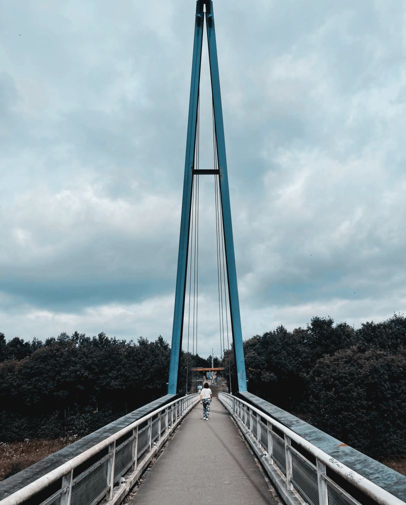 On se trouve sur un pont en plein milieu de la passerelle. On voit une personne de dos. À la fin du pont, deux poteaux de plusieurs mètres de haut forment un A.