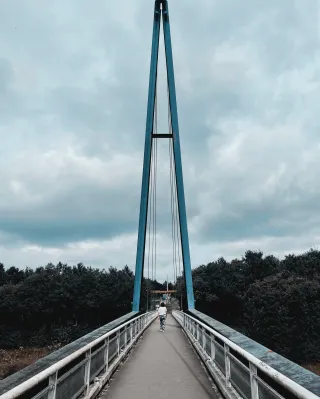 On se trouve sur un pont en plein milieu de la passerelle. On voit une personne de dos. À la fin du pont, deux poteaux de plusieurs mètres de haut forment un A.