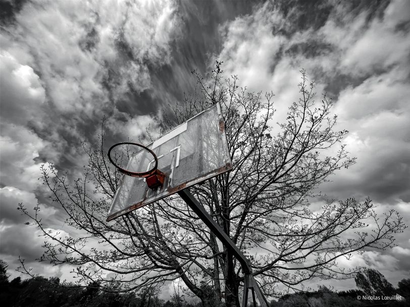 Photo en noir et blanc d'un panier de basket, vu du dessous. Seul l'arceau a gardé sa couleur rouge.
