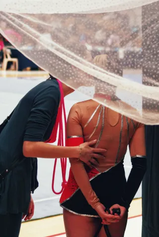 Deux personnes de dos. Celle de gauche parle à l'oreille de l'autre personne. Elle a sa main posée sur son dos. Les deux personnes sont derrière un voile blanc transparent. La personne de droite est en justaucorps, une tenue de GRS.