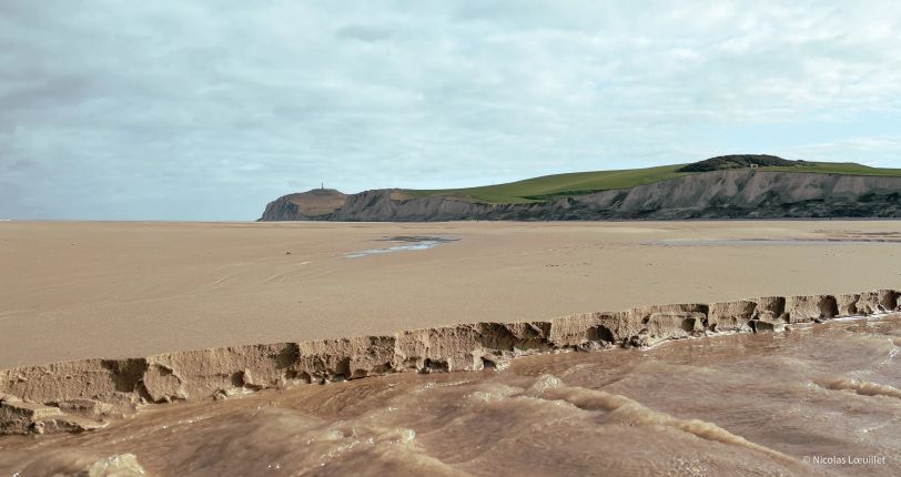 En arrière plan, la falaise du Cap Blanc-Nez. Au premier plan, l'eau a creusé le sable et donne l'impression qu'il y a une petite falaise de quelques centimètres de haut.