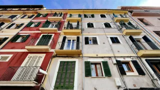 Une façade d'immeubles. On voit des balcons. Il y a un mur rouge, un mur crème. Les volets sont verts.