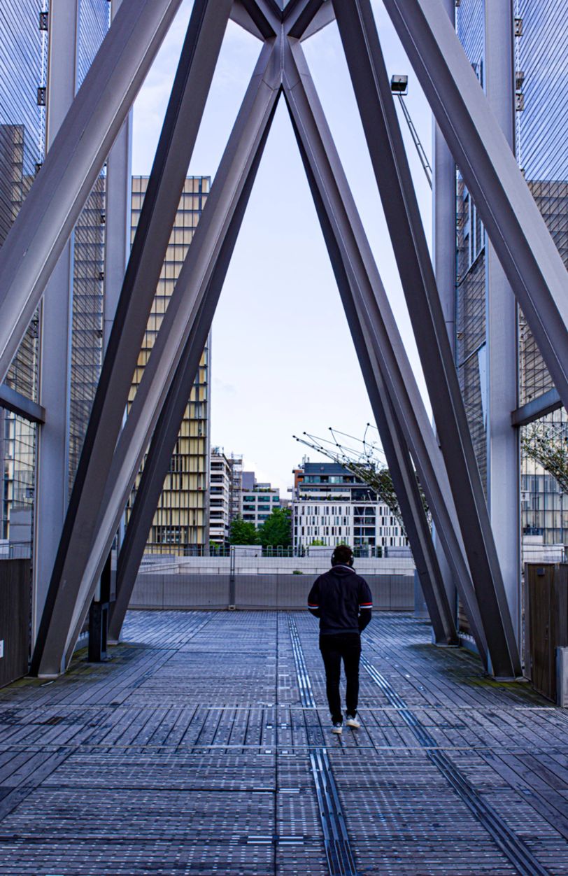 Une personne marche, on la voit de dos. Elle passe sous une structure métallique qui forme des grands compas.