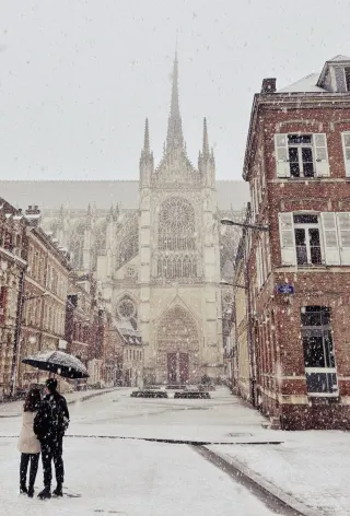 En arrière plan, on aperçoit la Cathédrale d'Amiens sous la neige. Au premier plan, un couple de dos, sous un parapluie, qui regarde la cathédrale.