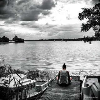 Une personne est assise sur un ponton. On la voit de dos. Devant elle, un lac.