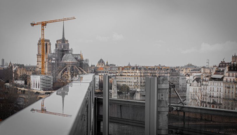 En arrière plan, on aperçoit Notre-Dame de Paris. En premier plan, on voit le reflet de Notre-Dame dans de l'eau stagnante sur une barrière.