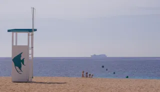 Photo d'une plage. La partie ensablée est légèrement surélevée par rapport à la mer. Entre le sable et la mer, on aperçoit un groupe de 4 personnes, en file indienne, de la plus grande à la plus petite. Comme les Dalton. Sur le sable, une cabine en bois. Dans la mer, un paquebot.