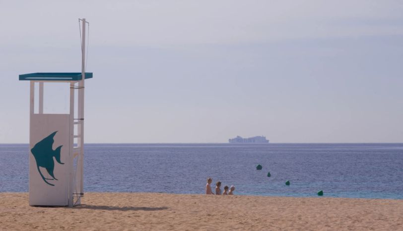 Photo d'une plage. La partie ensablée est légèrement surélevée par rapport à la mer. Entre le sable et la mer, on aperçoit un groupe de 4 personnes, en file indienne, de la plus grande à la plus petite. Comme les Dalton. Sur le sable, une cabine en bois. Dans la mer, un paquebot.