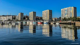Des immeubles qui se reflètent dans l'eau. Au premier plan, des bateaux de pêche.