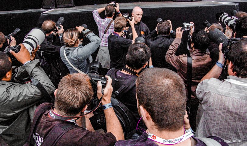 Photo prise durant un concert. Elle est cadrée sur un agent de sécurité, qui est entourée d'une dizaine de photographes qui prennent en photo la scène (qu'on ne voit pas).