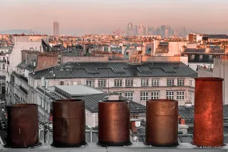 5 cheminées depuis une toiture. La plus grande se trouve à droite. On a l'impression que ce sont 5 personnes qui regardent les toits de Paris en arrière-plan.