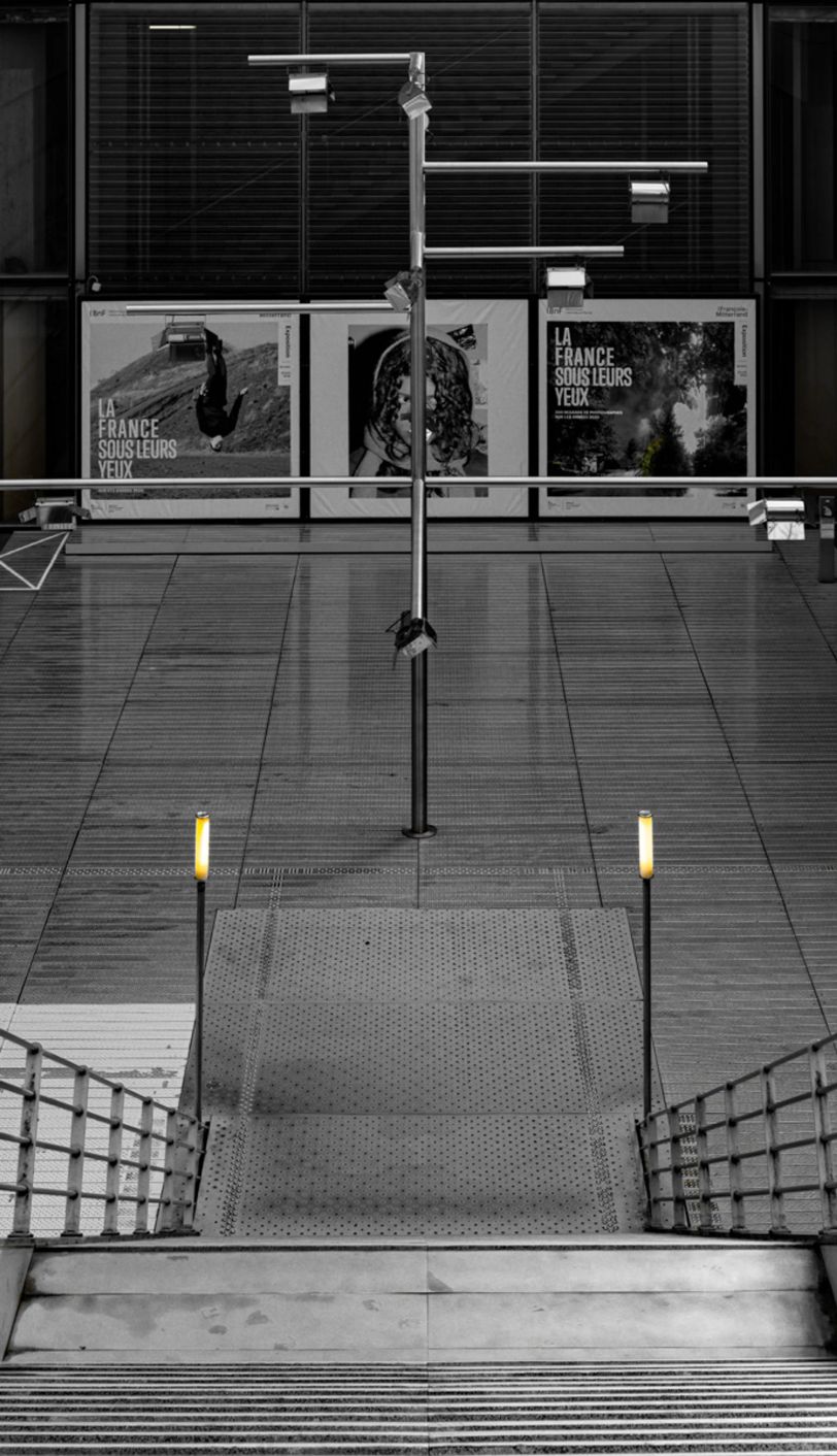 La photo est en noir et blanc. On se trouve en haut d'un escalier. Aux pieds de l'escalier, un lampadaire de part et d'autre. C'est la seule touche de couleur sur cette photo. Au fond, on aperçoit trois grandes affiches dont deux qui indiquent "La France sous leurs yeux".