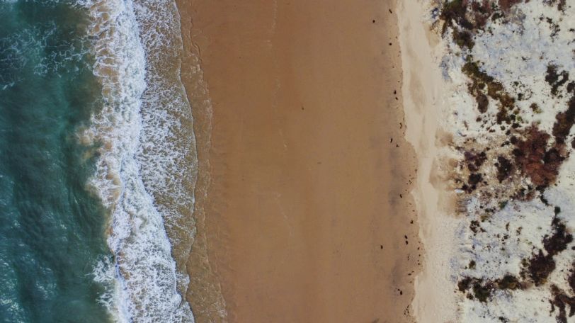 Photo d'une plage prise en drone, à la verticale. Un tiers de mer, un tiers de sable, un tiers de falaise.