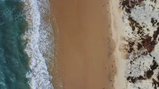 Photo d'une plage prise en drone, à la verticale. Un tiers de mer, un tiers de sable, un tiers de falaise.