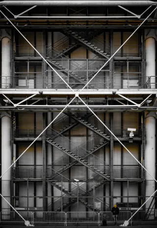 Au pied d'une structure métallique d'un bâtiment qui forme un X (on voit aussi des escaliers derrière ce X), une personne semble regarder son téléphone. Elle porte un bonnet jaune qui rappelle les reflets jaunes que l'on voit dans la structure métallique.