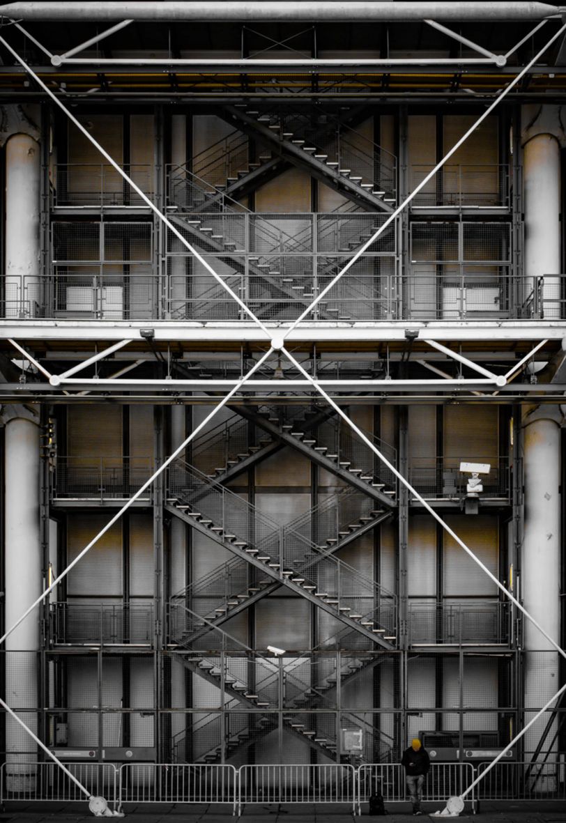 Au pied d'une structure métallique d'un bâtiment qui forme un X (on voit aussi des escaliers derrière ce X), une personne semble regarder son téléphone. Elle porte un bonnet jaune qui rappelle les reflets jaunes que l'on voit dans la structure métallique.
